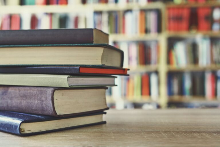 Stabel med bøker på et bord, med bokhyller i bakgrunnen.